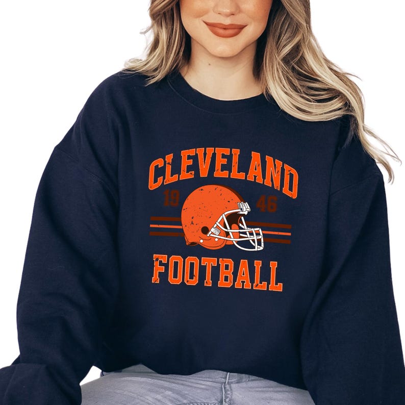May include: Navy blue sweatshirt with a vintage-style Cleveland Football logo featuring a brown and orange helmet with a distressed look. The text reads "CLEVELAND 1946 FOOTBALL".