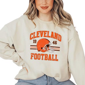 May include: A beige sweatshirt with a vintage-style graphic of a football helmet and the text "CLEVELAND FOOTBALL" with the numbers "19" and "46" on either side of the helmet.