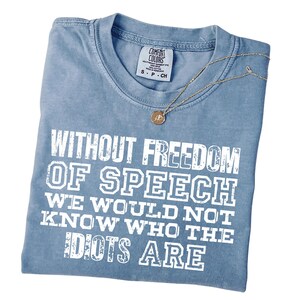 May include: A blue short-sleeved t-shirt with a distressed white graphic print that reads: WITHOUT FREEDOM OF SPEECH WE WOULD NOT KNOW WHO THE IDIOTS ARE. The shirt features a crew neckline and a Comfort Colors brand label at the inner collar.