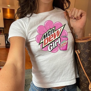 May include: White t-shirt featuring a graphic of a pink flower, a soda can, and the text "Mtn Dew Girl" in a stylized font. The shirt is a cropped style with short sleeves.