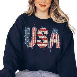 May include: A navy blue crewneck sweatshirt featuring the letters USA across the chest. The letters are designed with a distressed, frayed-edge fabric pattern that mimics the American flag, displaying red and white stripes and blue with white stars.