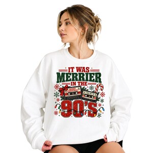 A woman wearing a white sweatshirt with a graphic design featuring Christmas-themed elements, including a cassette tape, snowflakes, and a ribbon.