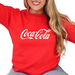 May include: Red sweatshirt with a white Coca-Cola logo printed on the front.