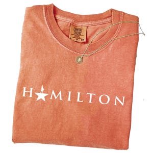 May include: Coral-colored t-shirt with the word "HAMILTON" in white block letters. A gold-colored necklace with a circular pendant rests on the shirt. The shirt has a Comfort Colors tag.