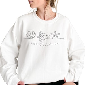 May include: White sweatshirt featuring a minimalist line drawing of a seashell, turtle, and starfish. Below the design is the text "Be still and know that I am God" and "Psalm 46:10" in black. The sweatshirt is a casual, comfortable style.