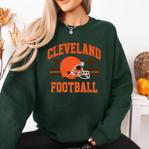 May include: A dark green sweatshirt with the text "CLEVELAND 19 46 FOOTBALL" and a brown and white football helmet graphic.