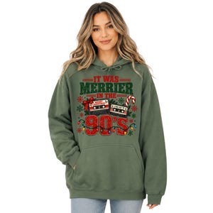 A woman wearing a green hoodie with a graphic design that says "IT WAS MERRIER IN THE 90'S" and features various Christmas-themed elements such as a snowflake, a candy cane, and a cassette tape.