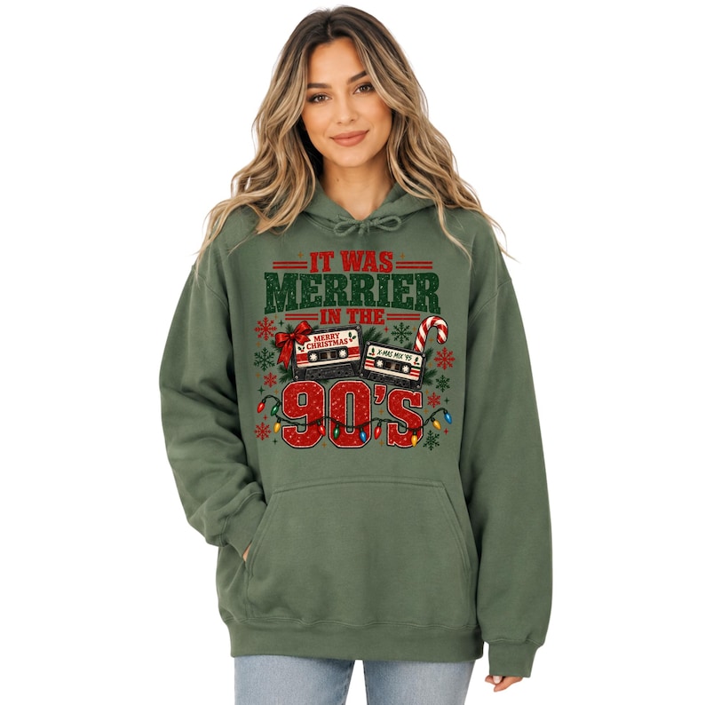 A woman wearing a green hoodie with a graphic design that says "IT WAS MERRIER IN THE 90'S" and features various Christmas-themed elements such as a snowflake, a candy cane, and a cassette tape.