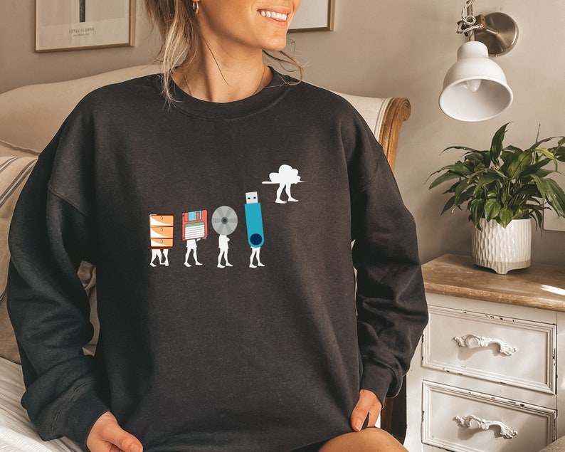 May include: Black sweatshirt with a graphic of a cloud carrying a USB drive, followed by a series of figures carrying different storage devices, including a floppy disk, a CD, and a hard drive. The text "The Evolution of Storage" is printed below the graphic.