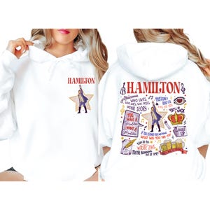 A white hoodie with a colorful graphic design featuring the character Hamilton from the musical Hamilton.