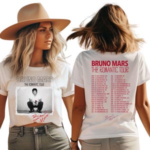 May include: White t-shirts featuring the text "BRUNO MARS THE ROMANTIC TOUR" in red. One shirt displays a photo of Bruno Mars with a rose. The other shirt lists tour dates. The shirts are worn by a person.