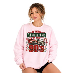 A woman wearing a pink sweatshirt with a graphic design that says "IT WAS MERRIER IN THE 90'S" and features various Christmas-themed elements such as a snowflake, a candy cane, and a radio.