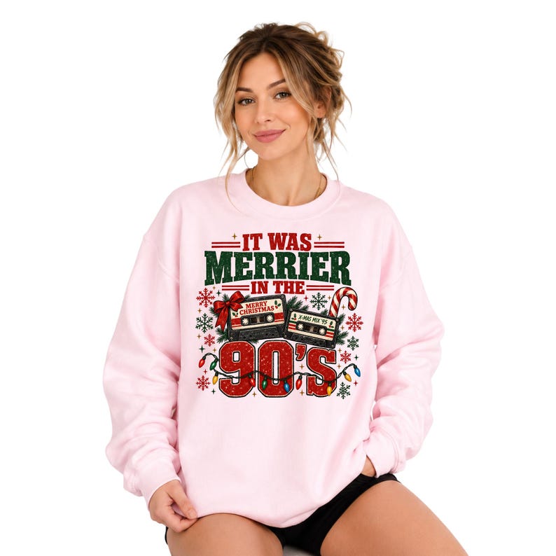 A woman wearing a pink sweatshirt with a graphic design that says "IT WAS MERRIER IN THE 90'S" and features various Christmas-themed elements such as a snowflake, a candy cane, and a radio.
