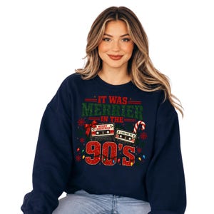 A woman wearing a navy blue sweatshirt with a graphic design featuring a Christmas tree, a cassette tape, and the text "It was merrier in the 90's".