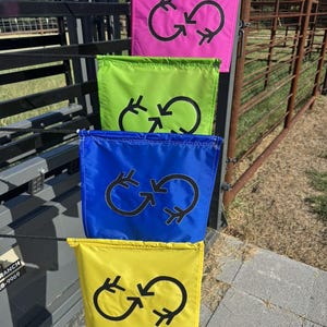 May include: Four brightly coloured flags in pink, green, blue, and yellow. Each flag features a black circular design with arrows. The flags are suspended from a black metal structure, with a brown metal fence visible. Likely used for signalling.