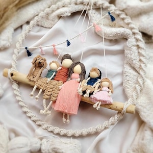 May include: A macrame wall hanging with a wooden dowel and six yarn dolls hanging from it. The dolls are dressed in different colors and have yarn hair. There are also small pink and blue yarn tassels hanging from the dowel.