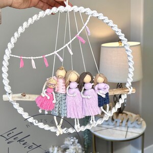 May include: A macrame wall hanging featuring five dolls with wooden heads and yarn hair, dressed in pink, gray, and blue outfits. The dolls hang from a driftwood shelf suspended within a white macrame circle, adorned with pink and white tassels.