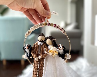 May include: A decorative hanging ornament featuring a bride and groom figurine with two small dogs on a wooden bar inside a gold hoop. The hoop is adorned with wooden letter beads spelling AUGUST 9 2025. The groom wears a brown suit, and the bride wears a white tulle dress.