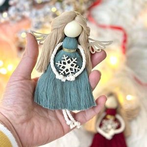 May include: A teal blue angel ornament with a white snowflake and gold trim. The angel has blonde hair and a wooden head. The ornament is held in a hand.
