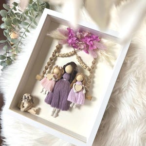 May include: A framed artwork featuring a family of three dolls on a swing, with a small dog figurine. The dolls wear purple and pink dresses and hold wooden hearts. The swing is adorned with the words "Summer" and "Autumn".