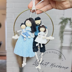 May include: A macrame family portrait with a gold hoop, a wooden dowel, and four doll-like figures. The figures include a woman in a light blue dress, a man in a blue sweater, and two children. The piece is suspended by a white cord.