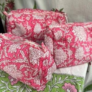 May include: Three pink cosmetic bags with white floral patterns. The bags are quilted and have a zip closure. The bags are different sizes and are arranged together. The background includes a green and white floral pattern.