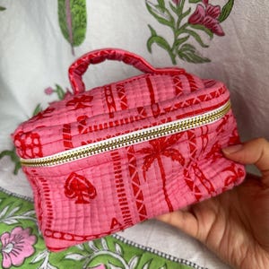 May include: A pink quilted cosmetic bag with a red pattern featuring playing card symbols and palm trees. The bag has a gold zip and a small handle. The bag is held in a hand.
