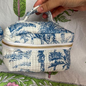 May include: A small, white cosmetic bag with a blue toile print featuring a tiger and foliage design. The bag has a gold zipper, a small handle, and a quilted texture. The bag is approximately 6 inches wide.
