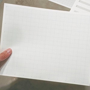 May include: A white grid paper sheet with a fine black grid pattern, held by a hand. Additional sheets with musical notation are visible in the background. The paper is designed for detailed drawings or diagrams.