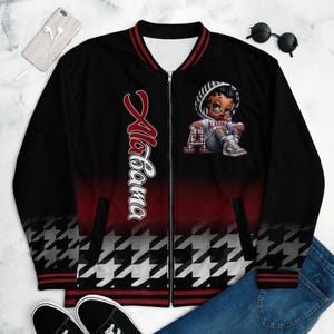 May include: Black bomber jacket with a red and white houndstooth pattern at the bottom. The jacket features the word "Alabama" in white script and a cartoon graphic of a character wearing a hoodie and sneakers.