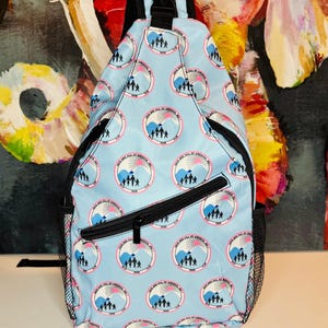May include: Light blue sling backpack with a repeating circular pattern featuring a mountain scene and the text "Jack and Jill of America, Inc. 1938". The bag has a black zipper, mesh side pocket, and adjustable strap.