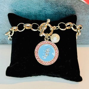 May include: A silver charm bracelet with a blue and pink charm featuring a Chinese character. The bracelet has a silver toggle clasp and a pearl charm.