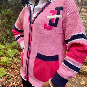 Puede incluir: Un c&aacute;rdigan rosa con cuello en V, con un dise&ntilde;o en contraste en blanco y negro en la parte delantera y las mangas. El su&eacute;ter tiene un bolsillo y est&aacute; adornado con un dise&ntilde;o de letras. Las mangas tienen rayas rosas, blancas y azul marino.