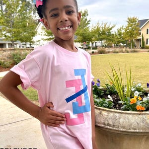 May include: A young person wearing a light pink t-shirt with a geometric design in blue and pink. The person is also wearing a large pink and blue bow in their hair. The word "SPLENDOR Bling" is visible at the bottom left.