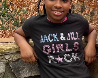 May include: A black t-shirt with the text "JACK & JILL GIRLS ROCK" in a sparkly, multi-colored design. The shirt has a pink collar and is worn by a young person. The background features a stone wall and foliage.