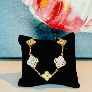 May include: Gold bracelet with four clover-shaped charms, each adorned with clear cubic zirconia stones. The bracelet is displayed on a black velvet cushion.