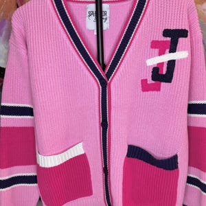 Puede incluir: C&aacute;rdigan rosa con ribetes azul marino y blancos, bolsillos y cierre de botones. El su&eacute;ter presenta las letras "TJ" en blanco, rosa y azul marino en la parte delantera. Las mangas tienen rayas rosas, azul marino y blancas. La marca "SPLENDOR Bling" es visible.
