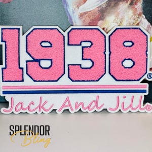 May include: Pink and white felt patch with the number 1938 in pink with a blue outline. The text "Jack And Jill" is written in pink cursive below the number. The patch has a white background and a blue and pink border.