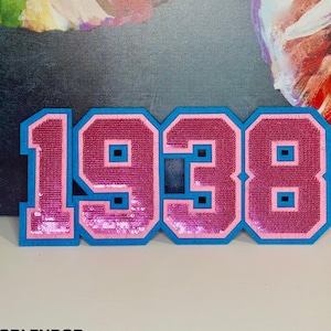 May include: Pink and blue sequined felt patch with the number 1938 in a varsity style font.