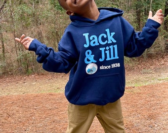 May include: A young person wearing a navy blue hoodie with the text "Jack & Jill" in light blue, paired with khaki pants and white sneakers. The hoodie also features a small patch with a mountain scene. The person is smiling and posing outdoors.