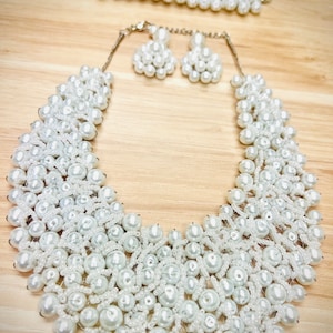 May include: A white beaded jewelry set featuring a necklace, bracelet, and earrings. The necklace is a bib style, and the earrings are small clusters. The bracelet and necklace have a silver chain clasp. The beads are various sizes.