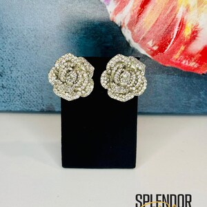 May include: A pair of silver rose-shaped earrings with clear rhinestones. The earrings are on a black display stand. The text "Splendor Bling" is visible below the stand.