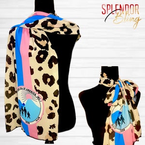 May include: A beige and brown leopard print scarf with a blue and pink stripe down the side. The scarf features a circular logo with the text "Land Till of America, Inc." and a graphic of a family standing in front of a mountain.