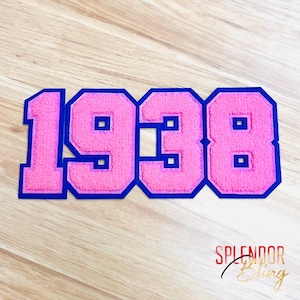May include: A pink, fuzzy patch with the number "1938" in a varsity style, outlined in blue. The patch is on a light wood-grain surface. The text "SPLENDOR BLING" is in the bottom right corner.