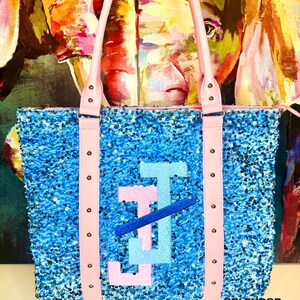 May include: A light blue tote bag with pink straps and a large blue, pink, and light blue letter "J" on the front. The bag is made of sequins and has a "Splendor Bling" logo on the bottom.
