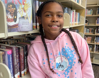 May include: A young person wearing a pink hoodie with a blue and pink heart design. The hoodie has a front pocket and a drawstring hood. The person is standing in a library next to a bookshelf with books.