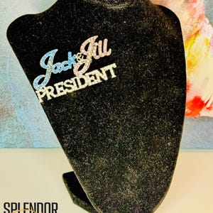 May include: A rhinestone brooch with the words "Jack & Jill PRESIDENT" in silver. The words "Jack" and "Jill" are in blue and pink rhinestones, respectively. The brooch is displayed on a black velvet bust.