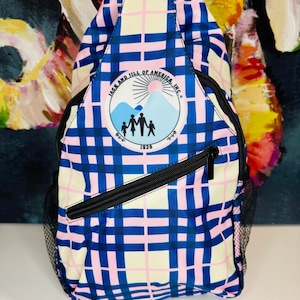 May include: A blue, pink, and cream plaid sling backpack with a black zipper and mesh side pocket. The backpack features a circular logo with a family silhouette and the text "JACK AND JILL OF AMERICA, INC." and "1938". The text "SPLENDOR Bling" is also visible.