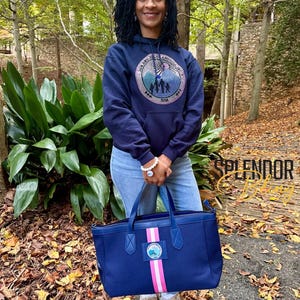 May include: A navy blue tote bag with a pink and white stripe and a circular emblem. The bag has a matching navy blue handle and a zipper closure. The bag is held by a person wearing a navy blue hoodie with a circular emblem.