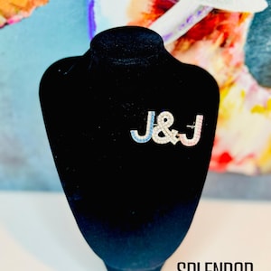 Pu&ograve; includere: Spilla color argento con le lettere "J & J" in blu e rosa, impreziosita da cristalli trasparenti. La spilla &egrave; esposta su un busto di velluto nero. Il testo "SPLENDOR Bling" &egrave; visibile in basso.